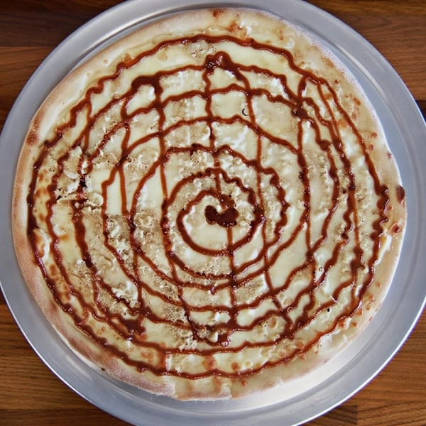 Dessert pizza with white frosting and a spiral of chocolate sauce on a metal tray.