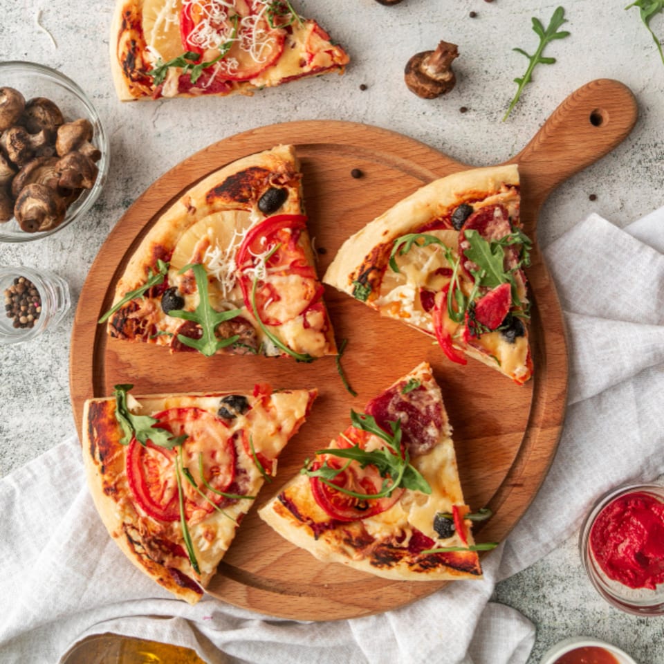 Greco's Pizzeria's sliced pizza with various toppings, including pepperoni, olives, and arugula, on a wooden board.